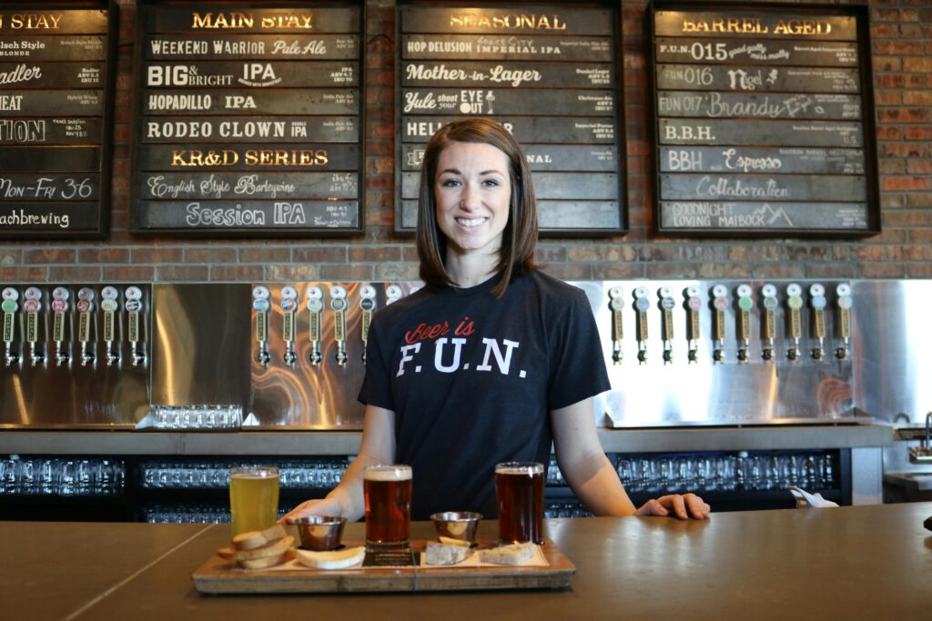 Smiling bartender in a bar serving craft beer flights with taps and menu board in the background.