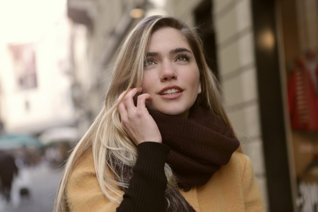 Close-up of a smiling woman outdoors with a scarf and brown coat, looking up.