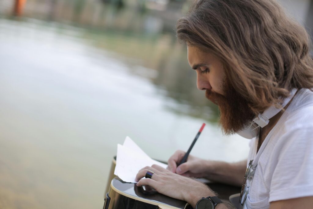 pexels-photo-3780003-3780003 An adult man with a guitar writes notes while relaxing outside by the lake.