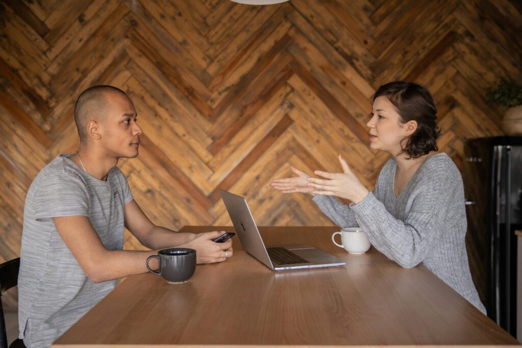 pexels-photo-4049960-4049960 Focused woman explaining opinion to ethnic male coworker during business teamwork sitting at table with laptop and coffee cups in cozy kitchen against wooden wall and looking at each other