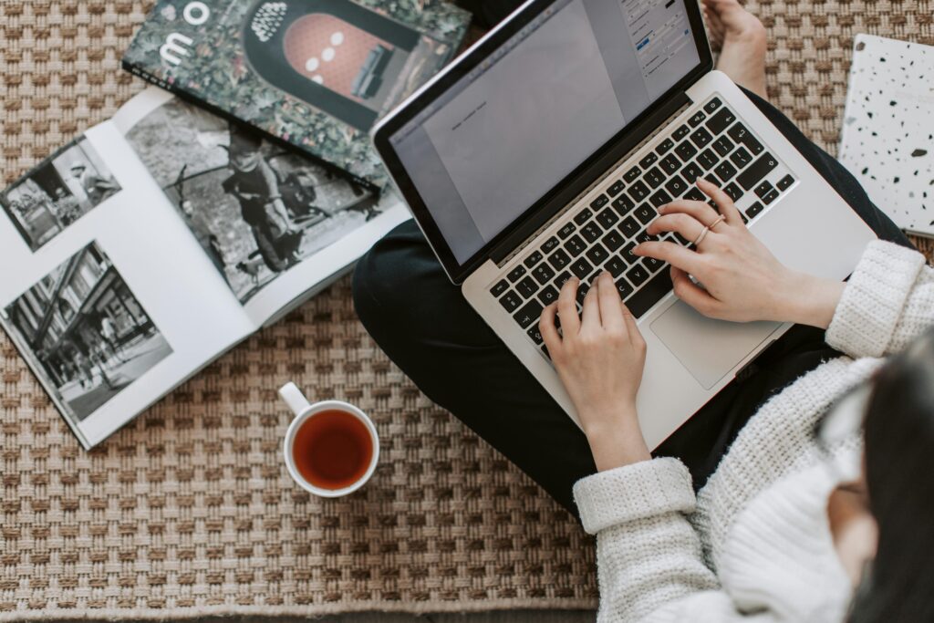 pexels-photo-4050347-4050347 Overhead view of a woman typing on a laptop at home, surrounded by magazines and a cup of tea on the floor.
