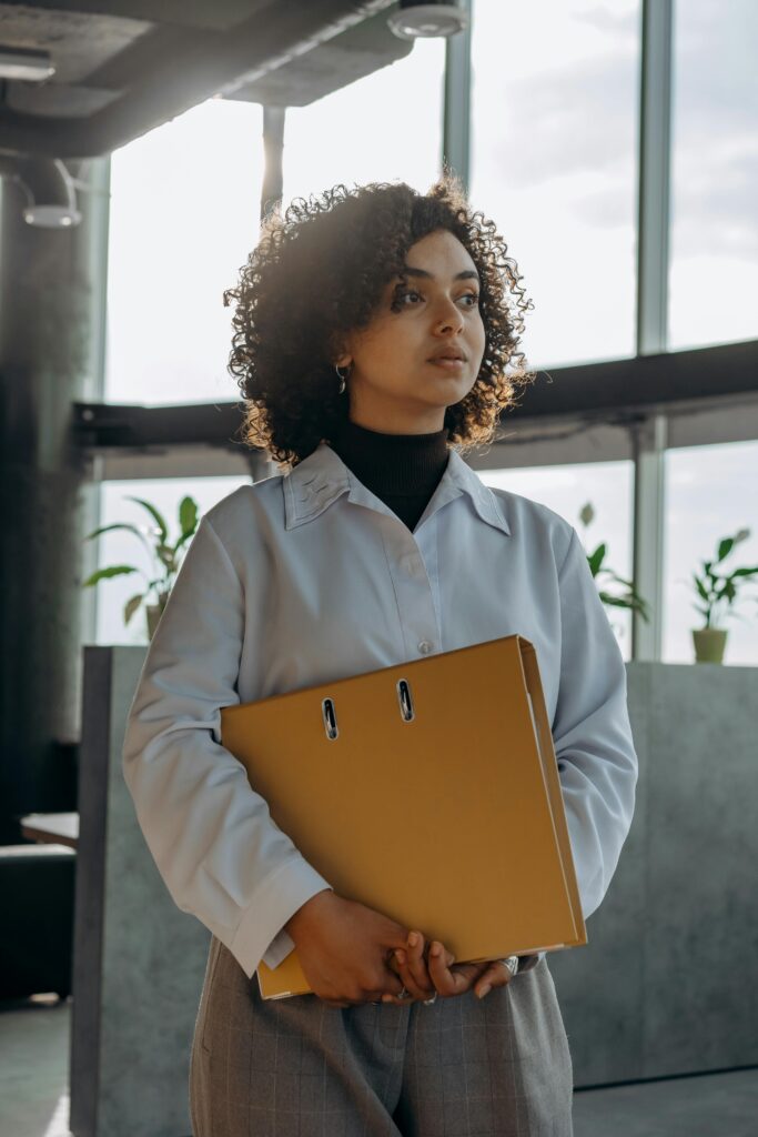 pexels-photo-8297138-8297138 A woman in professional attire holds a folder in a well-lit modern office setting.